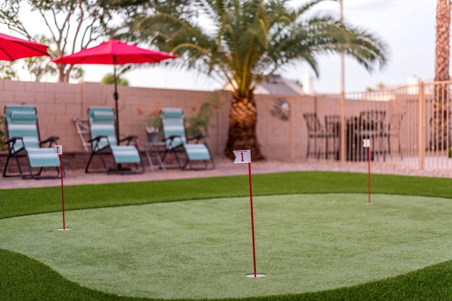 A small putting green with three numbered red flags in a backyard setting, crafted by expert artificial grass installers. The area is surrounded by lounge chairs with colorful cushions, red patio umbrellas, a palm tree, and a fenced area with a patio table and chairs in the background. Free estimates available for turf installations Houston.