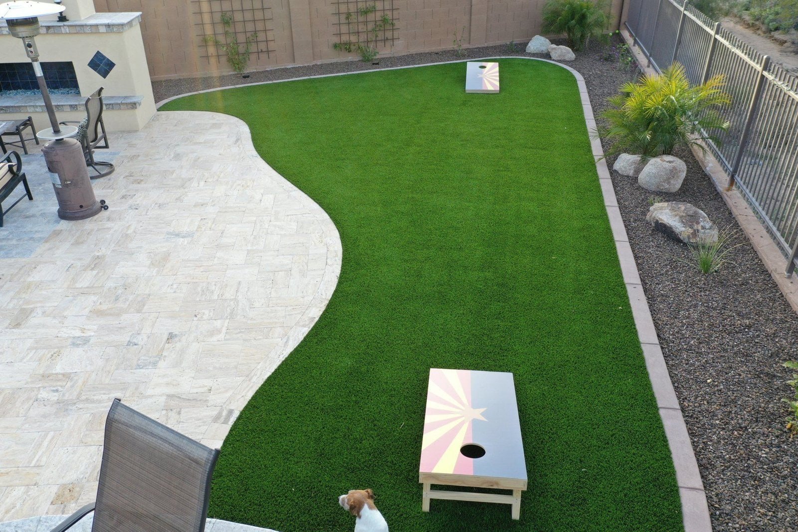 A backyard in Houston, TX, boasts a lush artificial turf lawn with two cornhole boards. There is a paved patio area adorned with chairs, a fire pit, and other outdoor furniture. A small dog sits at the edge of the turf, while pristine landscaping is enclosed by a fence and stone wall.