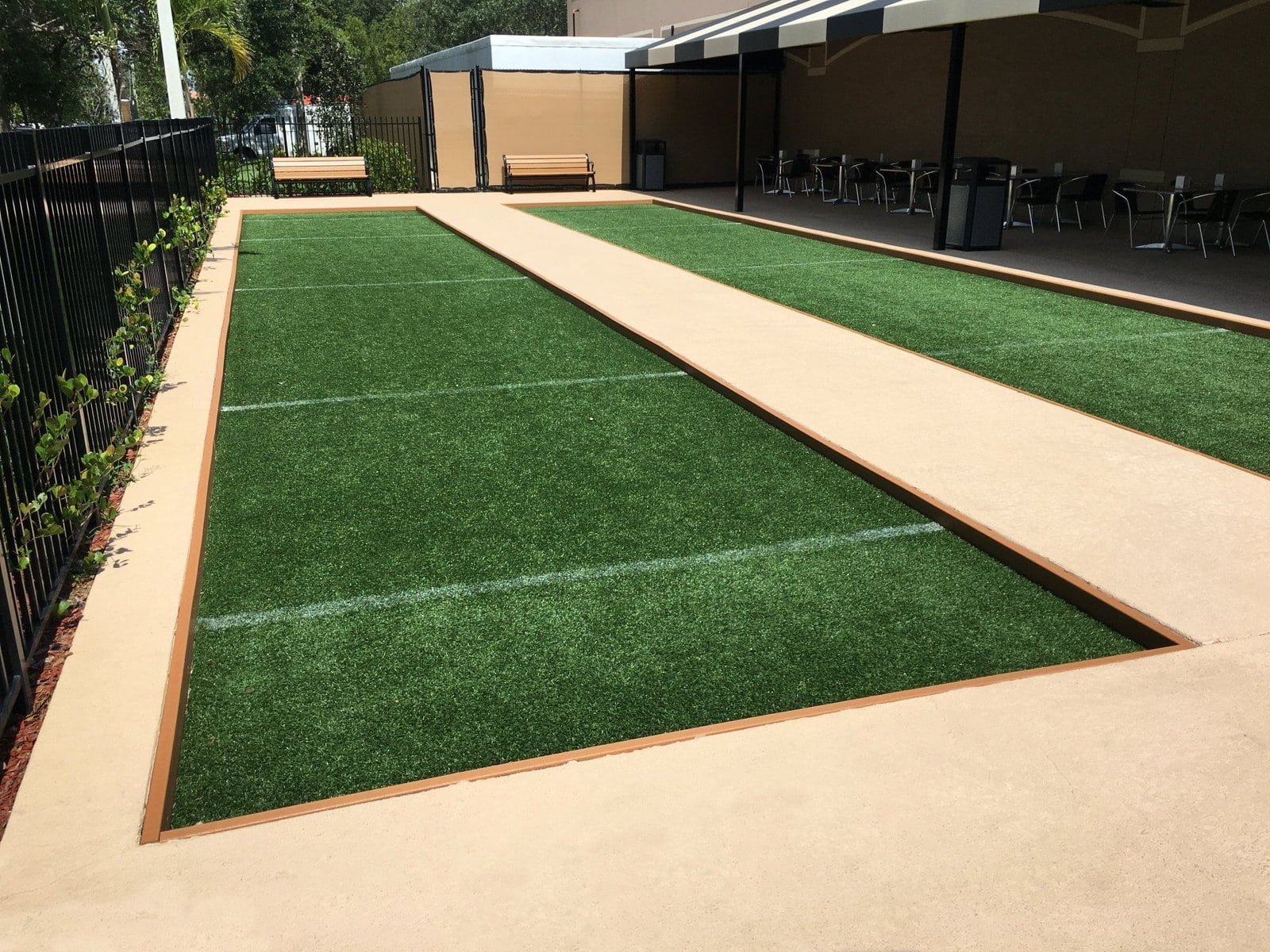 A well-maintained outdoor bocce ball court with green sports turf set between concrete borders, expertly installed by our dedicated installation team. A bench is visible at the end of the court, and to the right is a shaded area with tables and chairs. The area is enclosed by a metal fence with climbing plants.