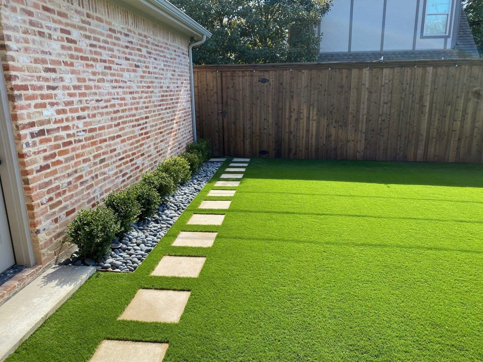 A backyard in Houston TX with a neatly manicured lawn, featuring evenly spaced beige stepping stones leading to a wooden fence. To the left of the stones, a row of small bushes and gray decorative rocks line the brick exterior of a house, showcasing expert turf installations by Artificial Grass Installers.