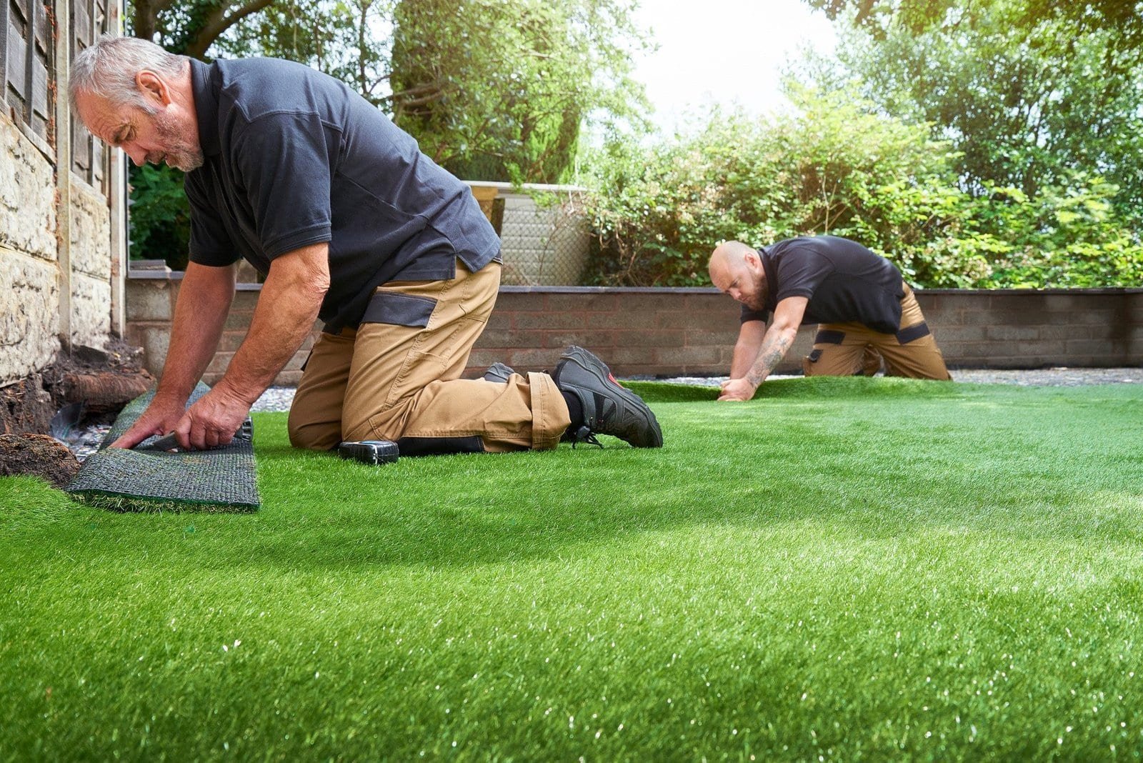 Two men kneeling on a lawn in Houston, TX, are installing artificial turf for a putting green in a garden. Focused on aligning and securing the turf near a house, both are dressed in black shirts and khaki pants. The garden is beautifully surrounded by greenery and trees in the background.